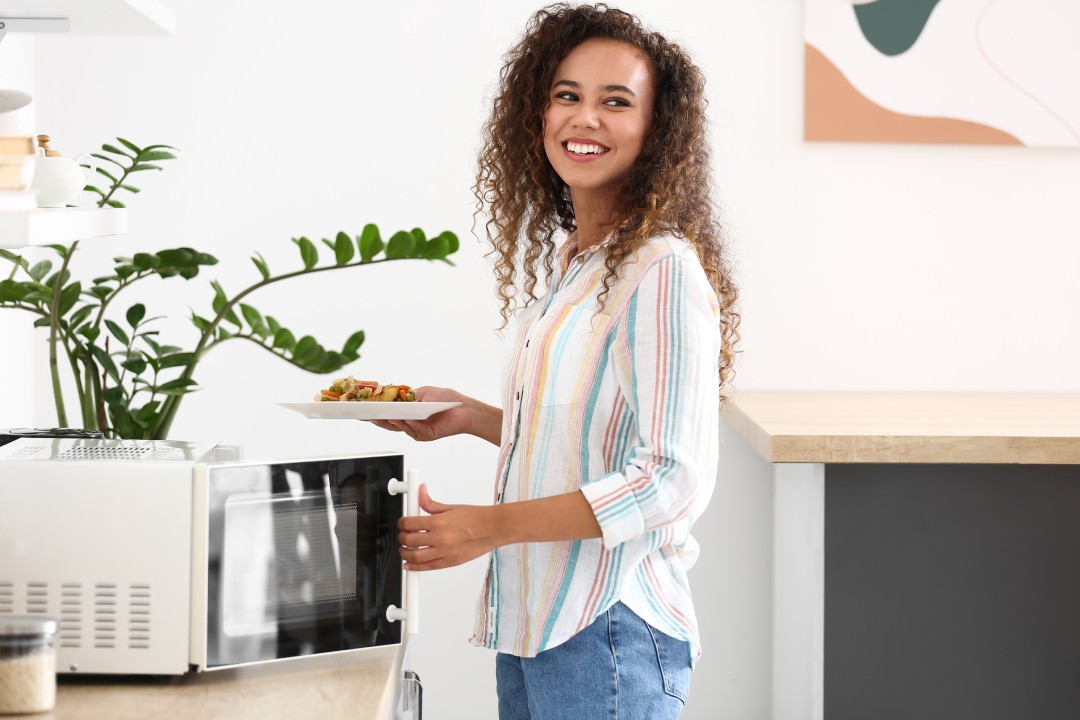 Woman putting plate with food into microwave oven in kitchen